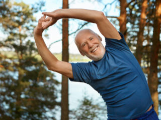 Outdoor shot of happy energetic senior retired man enjoying physical training in park, doing side bends exercise, holding hands together, looking at camera with broad smile, warming up body before run